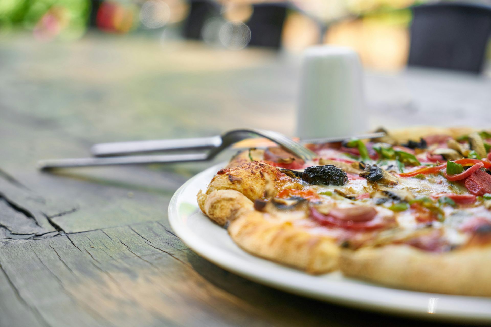 Pizza on a white plate with cutlery on a weathered wooden table outdoors.
