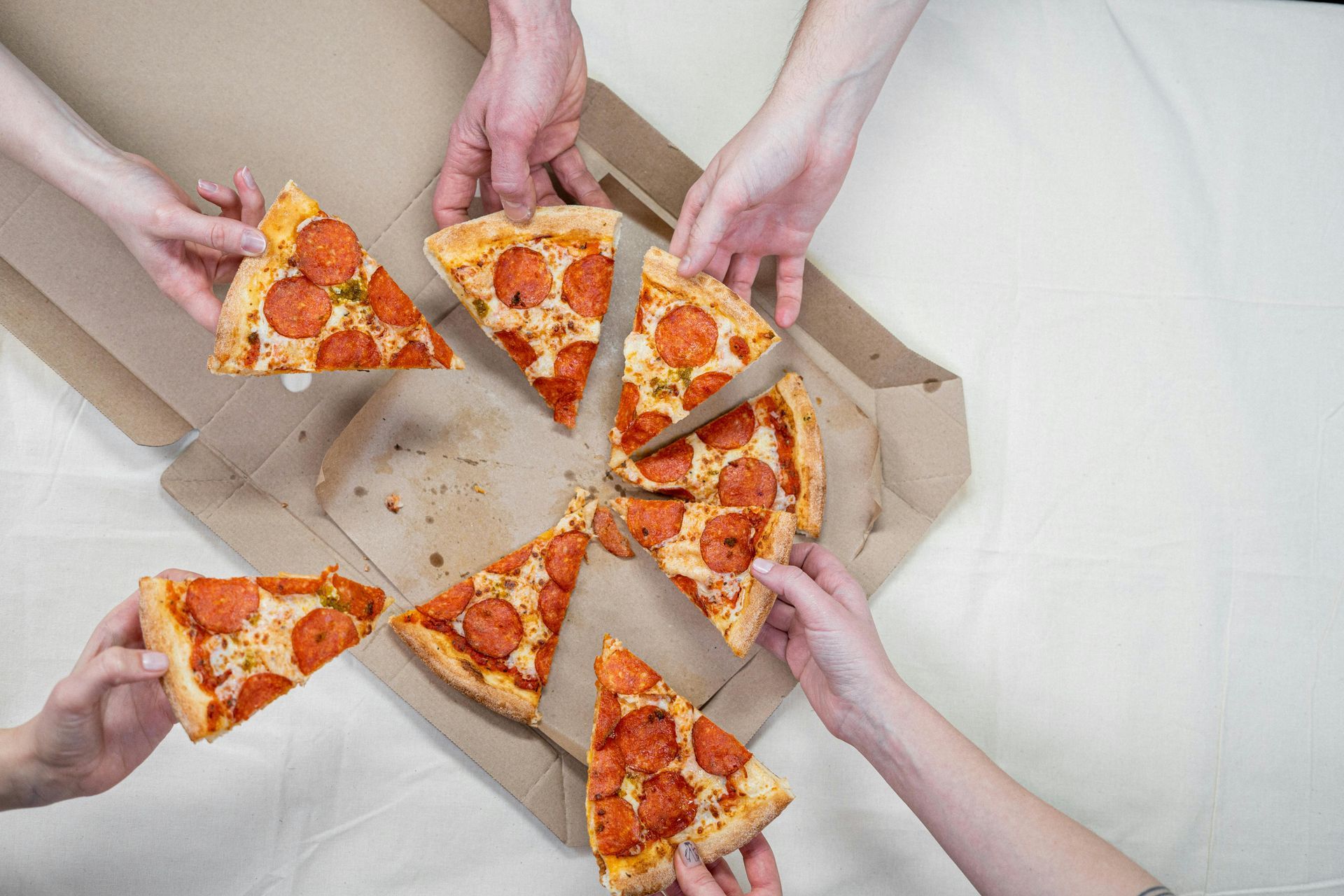 Hands reaching for pepperoni pizza slices from an open box on a white surface.