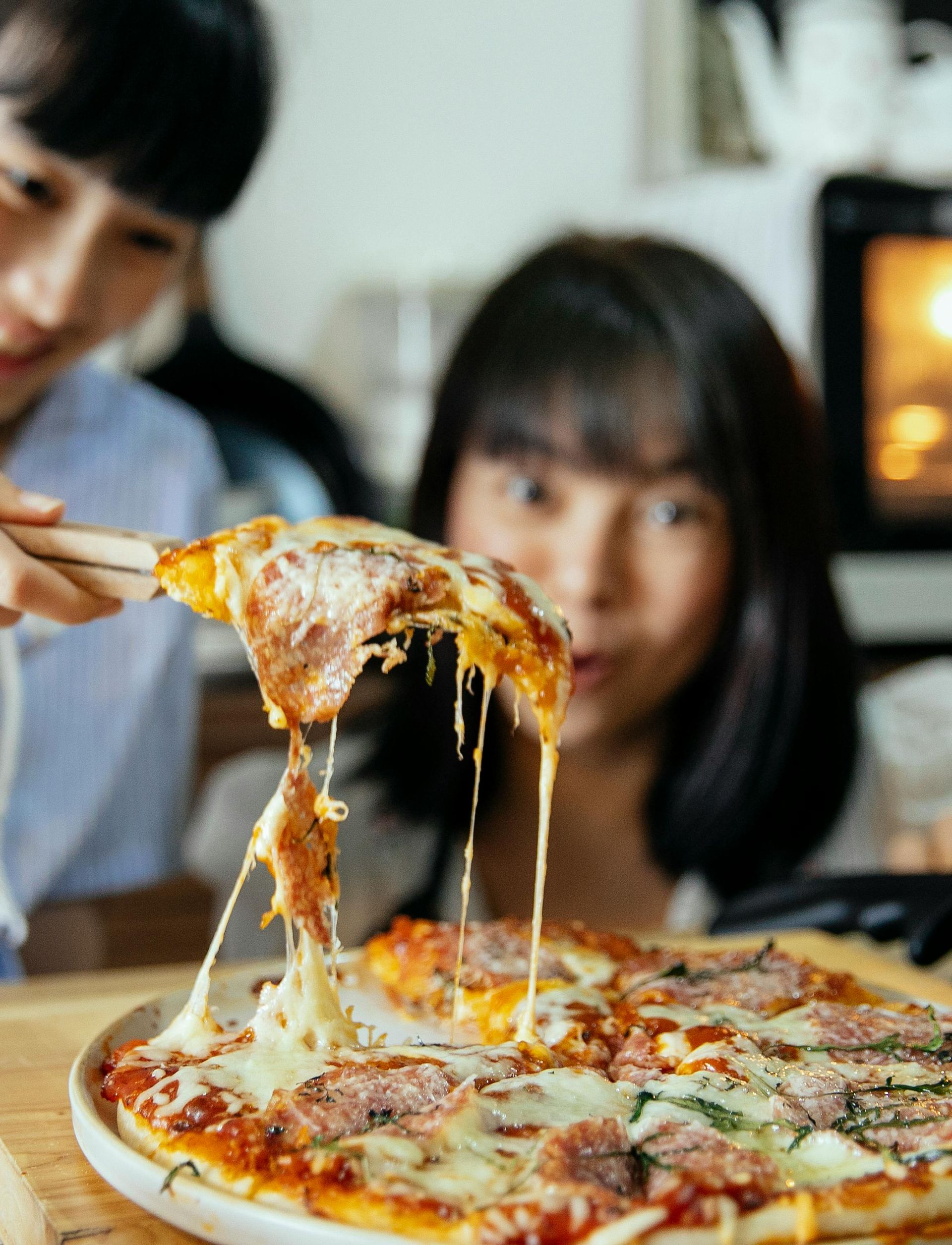 Two people look at pizza with long, stringy cheese. One person holds a pizza slice with chopsticks.