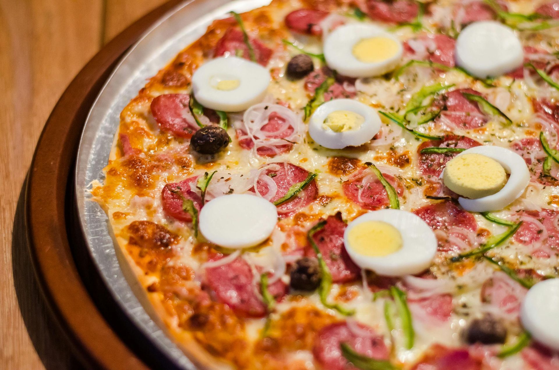 Pizza with sausage, sliced eggs, olives, onions, and green peppers on a metal tray.