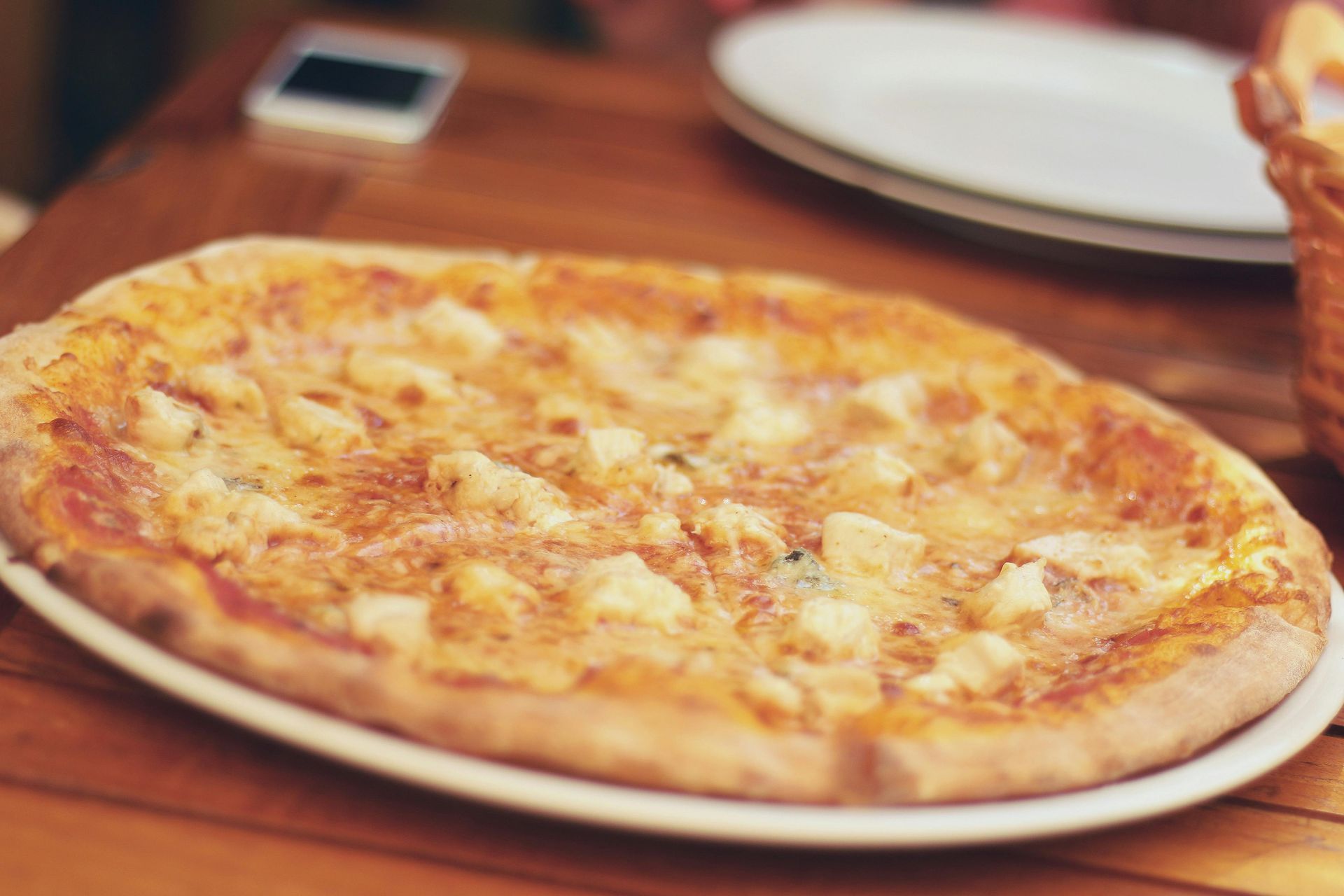 Pizza on a white plate on a wooden table. A phone and another plate are also on the table.