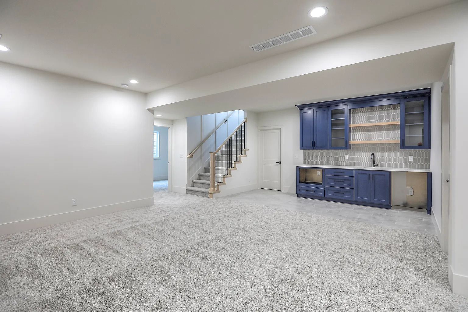 Empty basement room with grey carpet, blue bar area, and staircase.