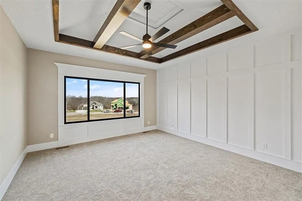 Empty room with beige walls, white paneling, large window, and carpet. Dark wood ceiling beams and a ceiling fan.