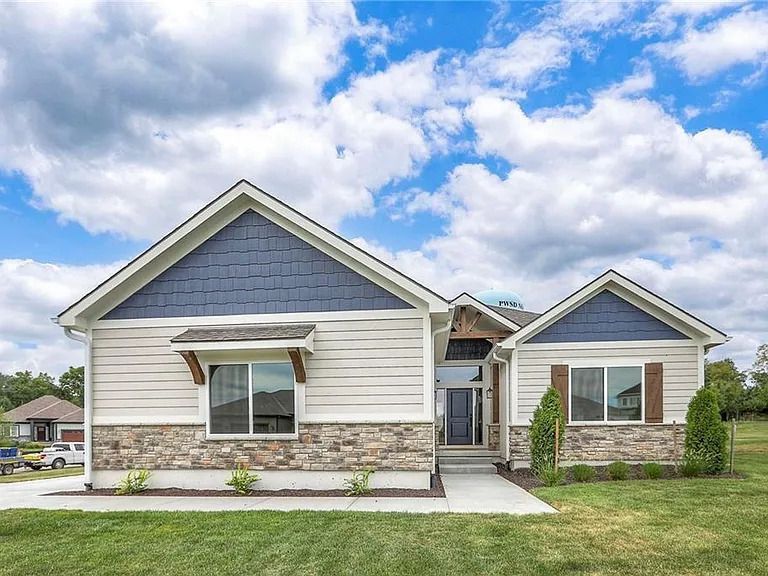Ranch-style house with tan siding, blue accents, stone facade, and a well-kept lawn under a cloudy sky.