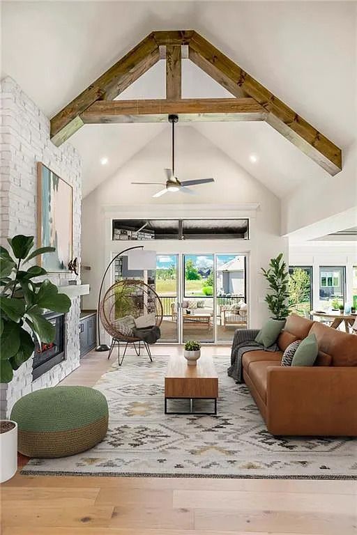 Spacious living room with vaulted ceiling, exposed beams, fireplace, leather sofa, and large windows overlooking a backyard.