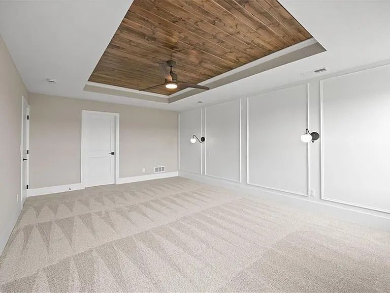 Empty bedroom with light carpet, white walls with panels, wood ceiling detail, and a ceiling fan.