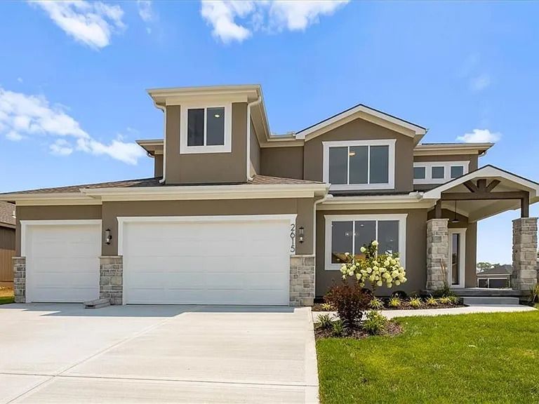 Two-story beige house with white garage doors, stone accents, and a blue sky.