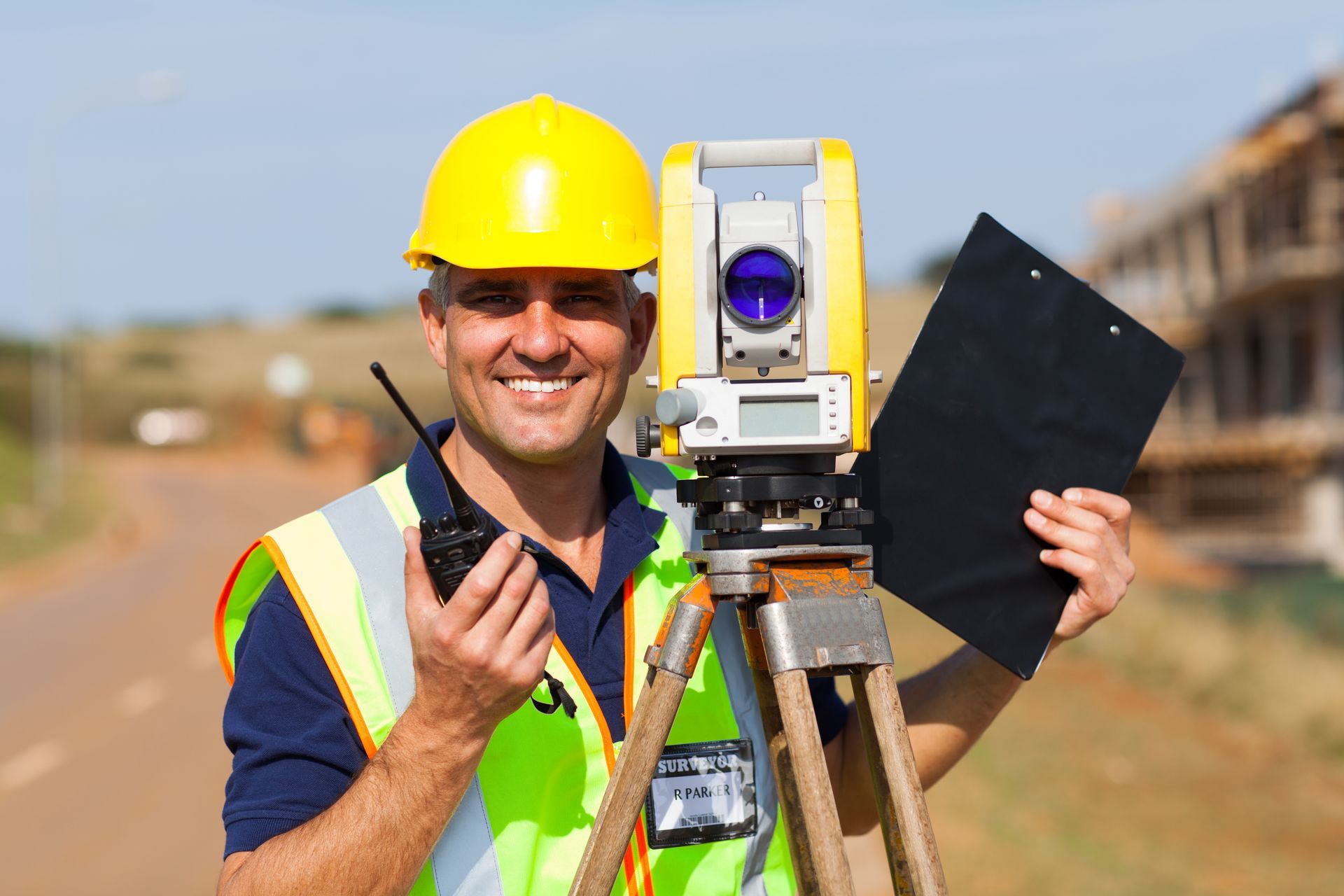 Smiling surveyor wearing a hard hat and safety vest, holding a radio and clipboard near surveying equipment on a road.