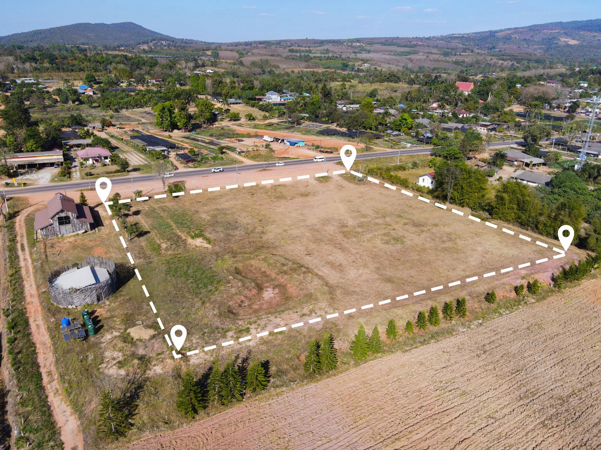 Aerial view of a rectangular vacant lot with four markers, surrounded by trees, a road, and rural buildings.