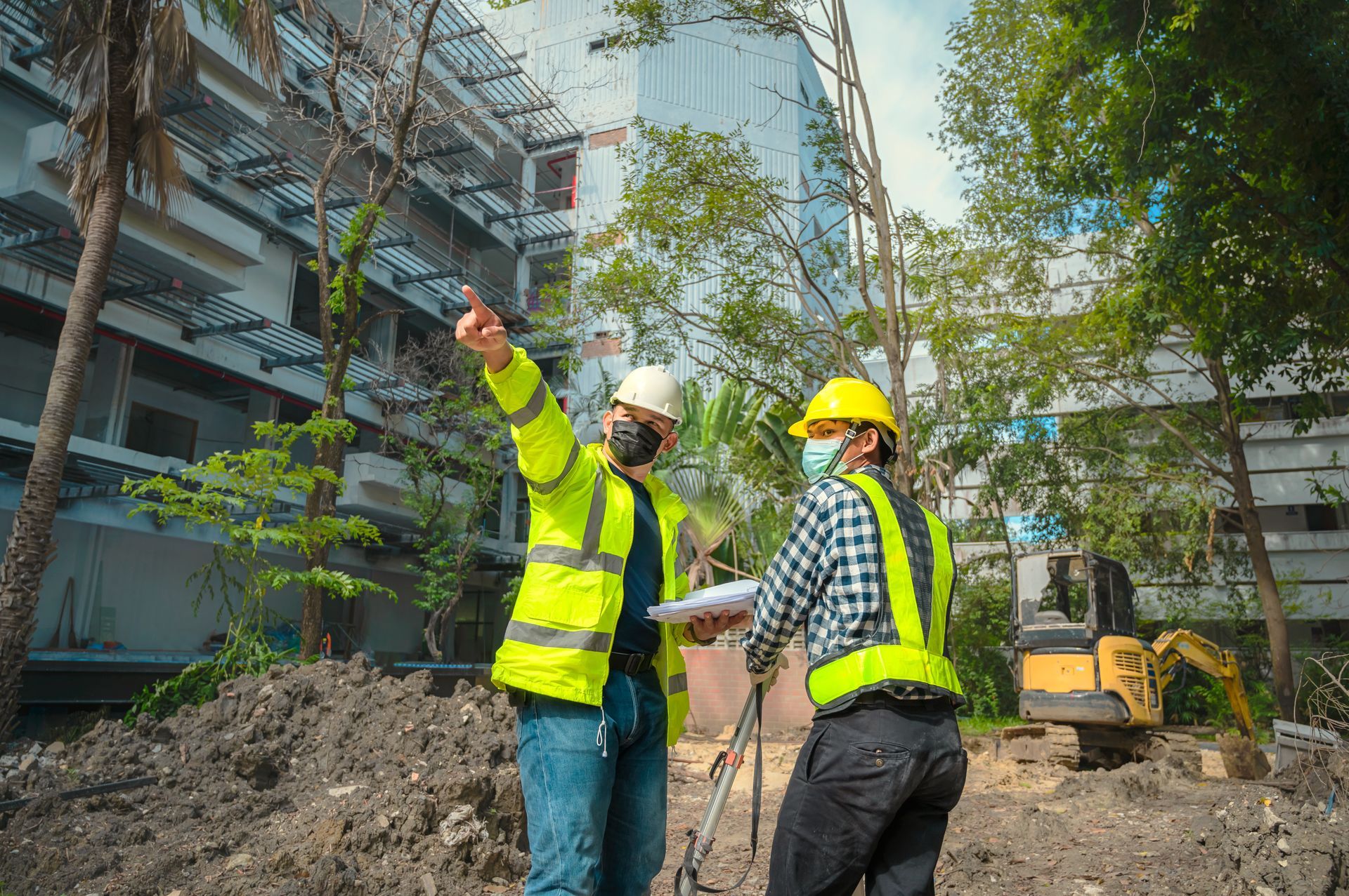Two construction workers in safety vests and helmets, reviewing plans at a construction site.