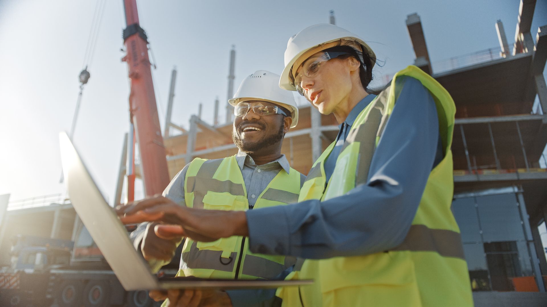 Two construction workers in safety vests and hard hats looking at a laptop at a construction site.