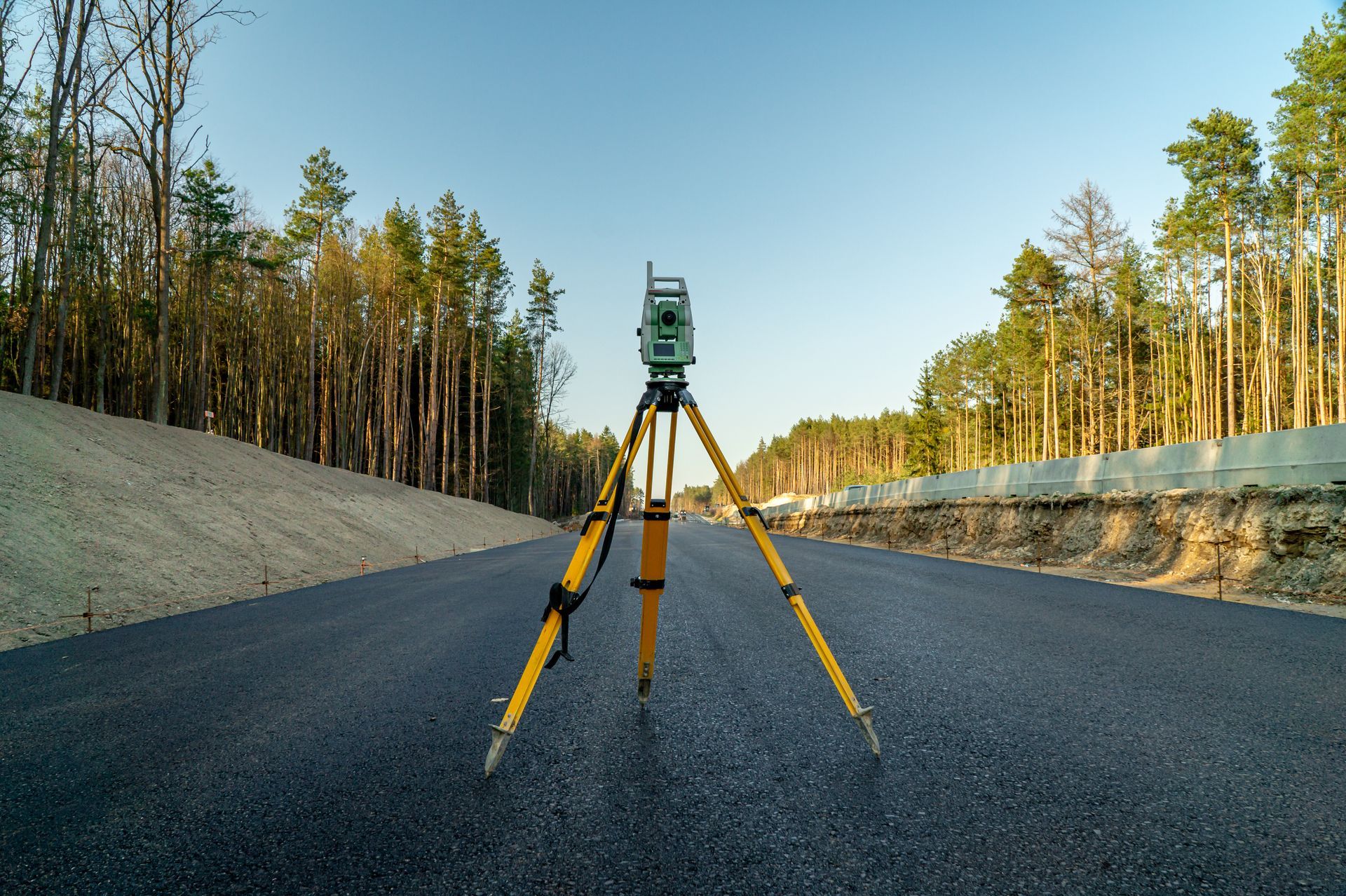 Surveying equipment on a freshly paved road, flanked by trees and a clear blue sky.