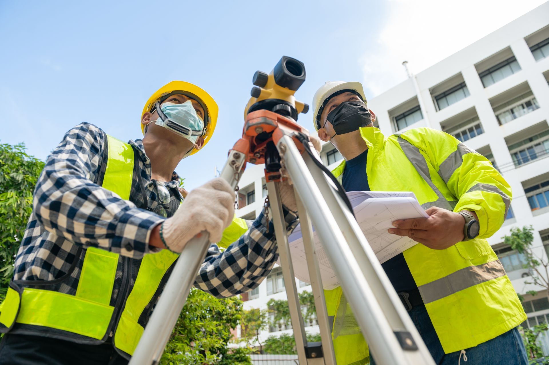 Two construction workers using surveying equipment outdoors, wearing masks and safety vests.