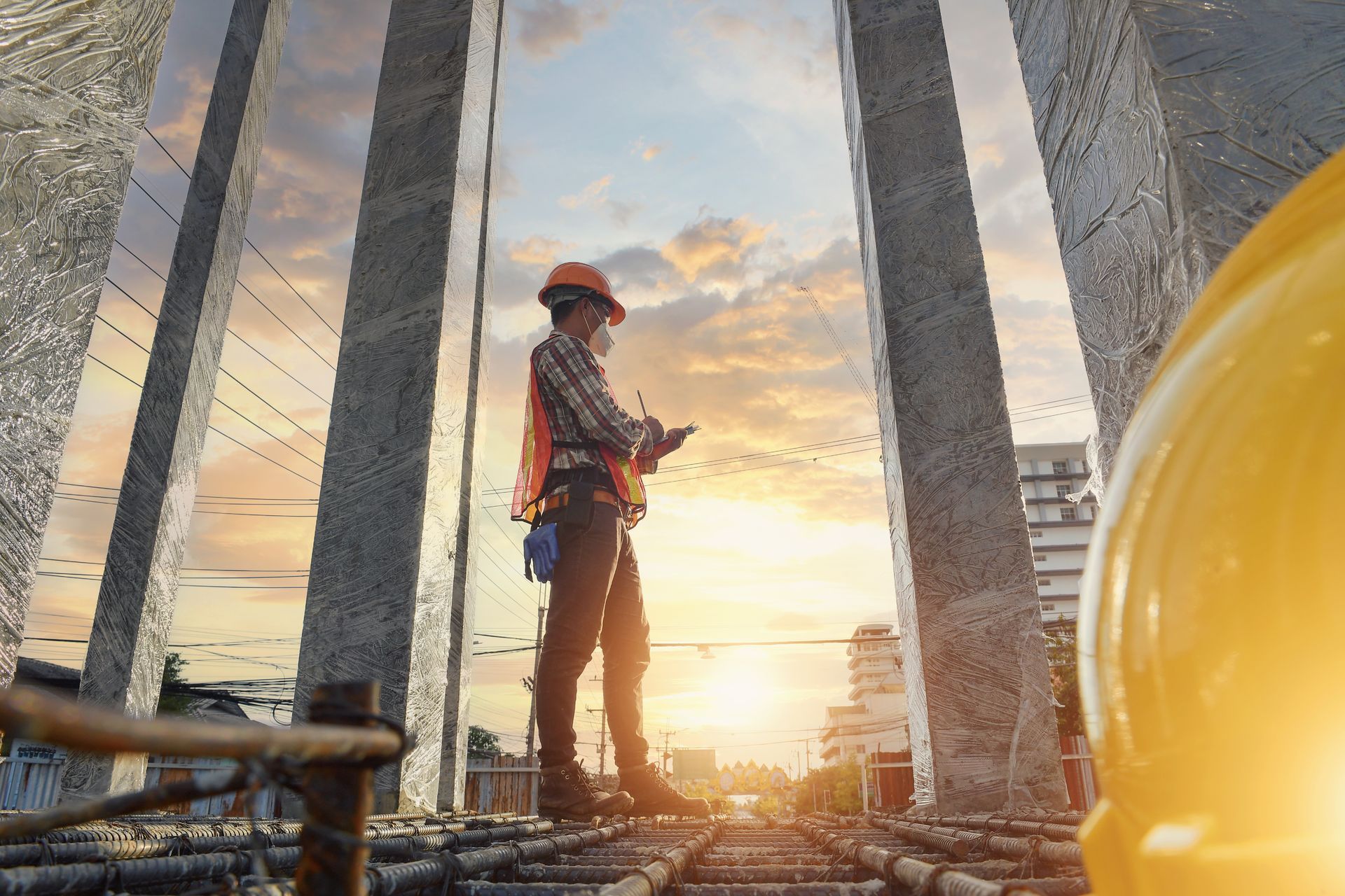 Construction worker, helmet and vest, standing among concrete pillars, overlooking a construction site at sunset.