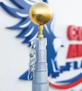 Close-up of a silver flagpole topped with a gold ball finial, with a chain and blue and red logo in the blurred background.