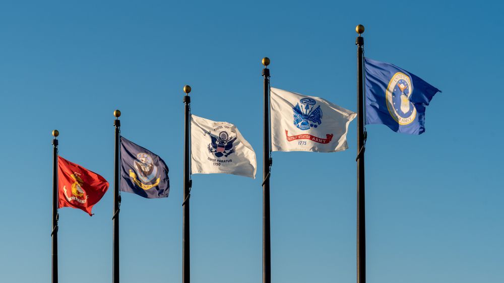 Flags of the U.S. Marine Corps, Navy, Army, Air Force, and Coast Guard fly against a blue sky.