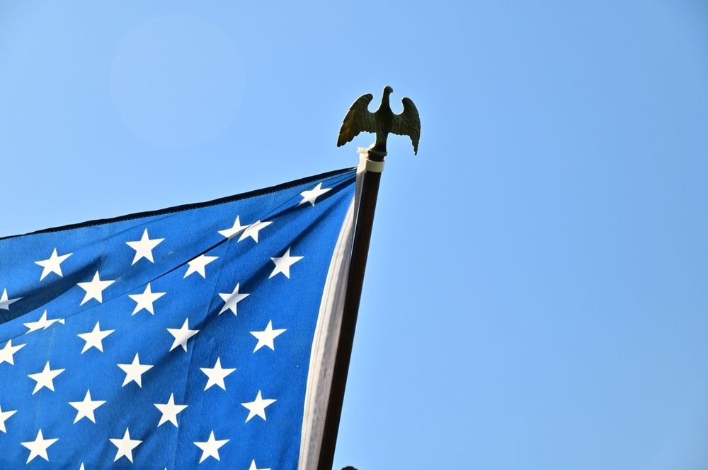 American flag with stars, blue field, and golden eagle on flagpole against a blue sky.