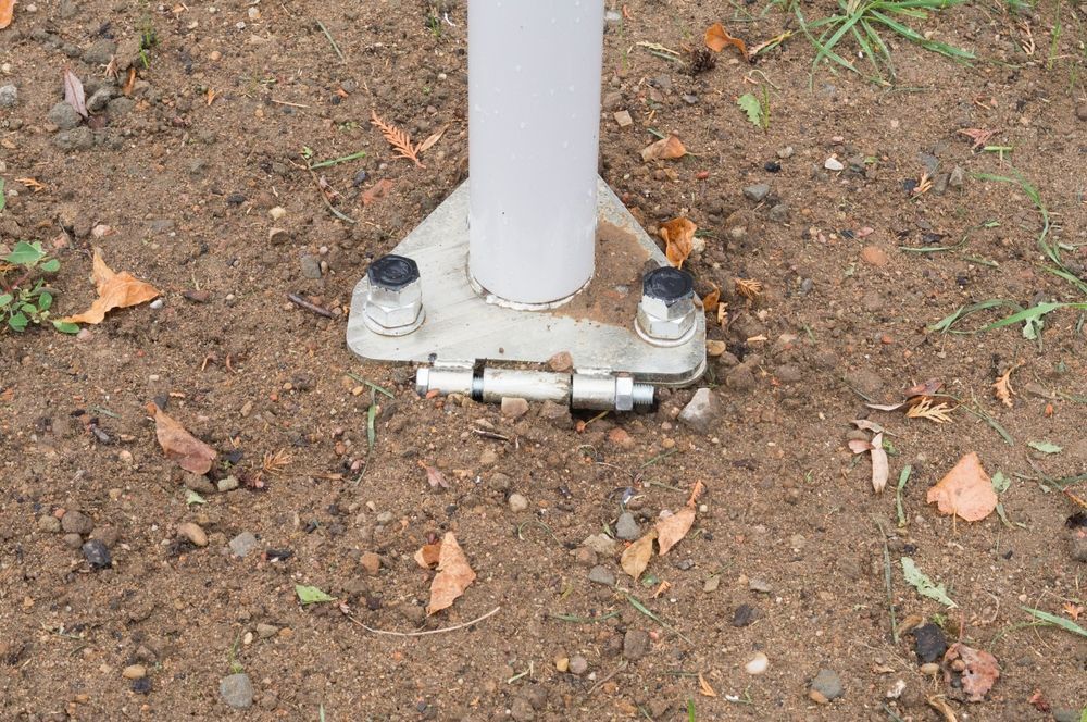 Base of a white pole secured to the ground with metal bolts on a metal plate, set in dirt.