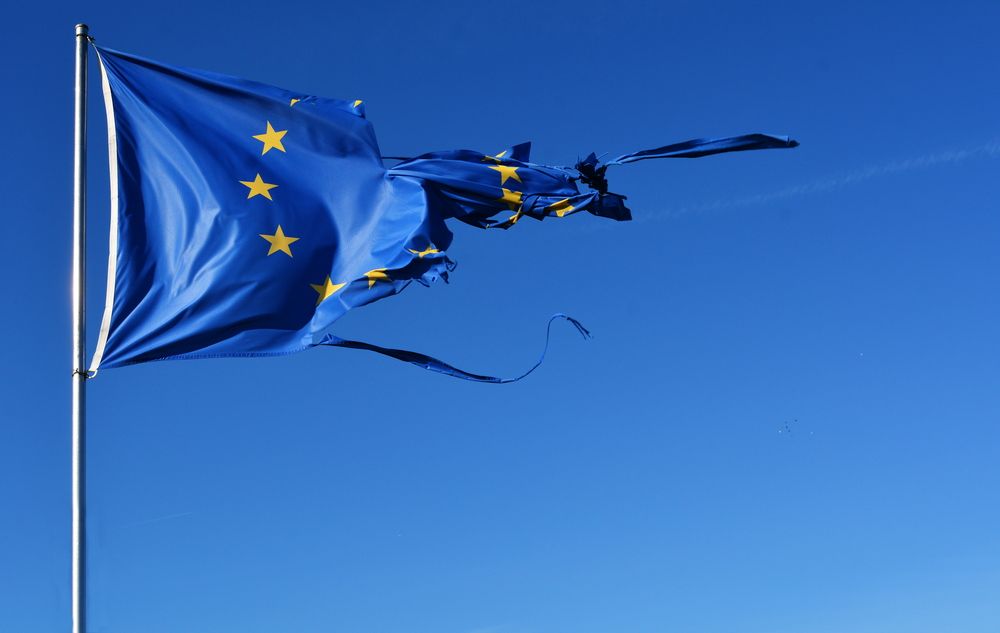 European Union flag, torn and tattered, flies against a bright blue sky.