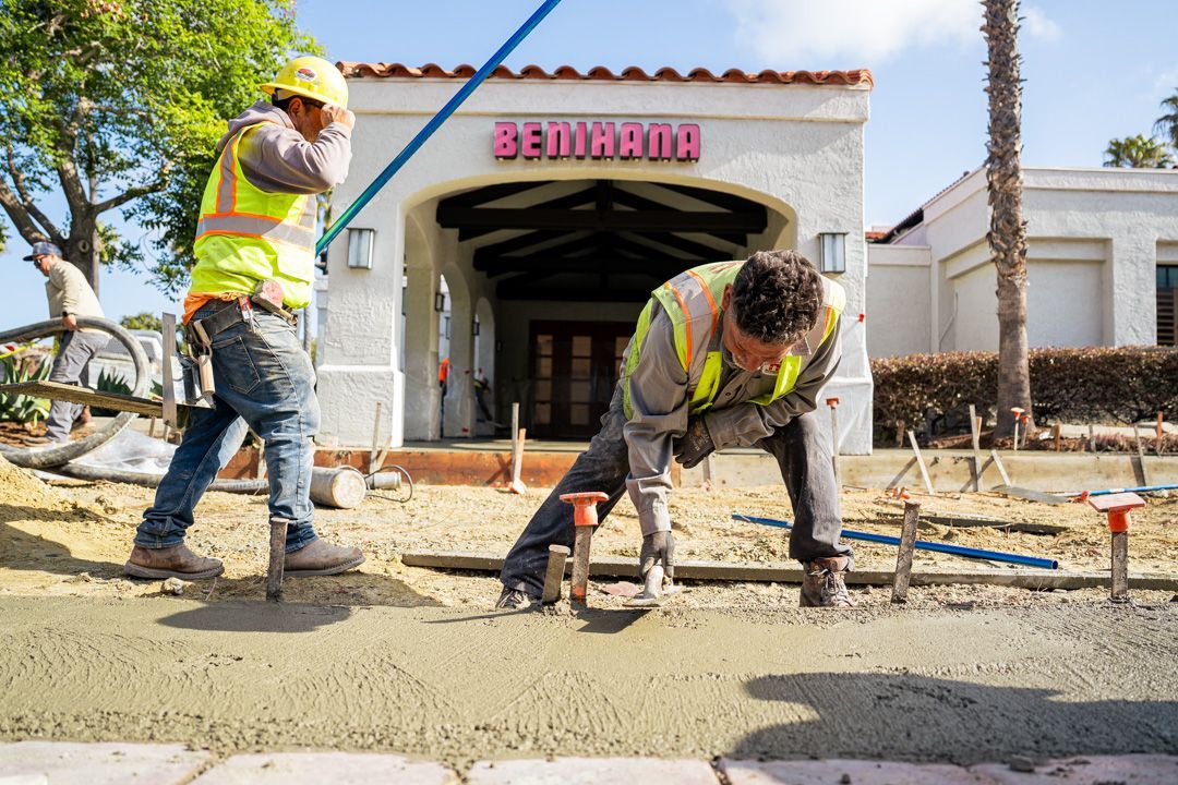 Two construction workers are working on a sidewalk in front of a building.