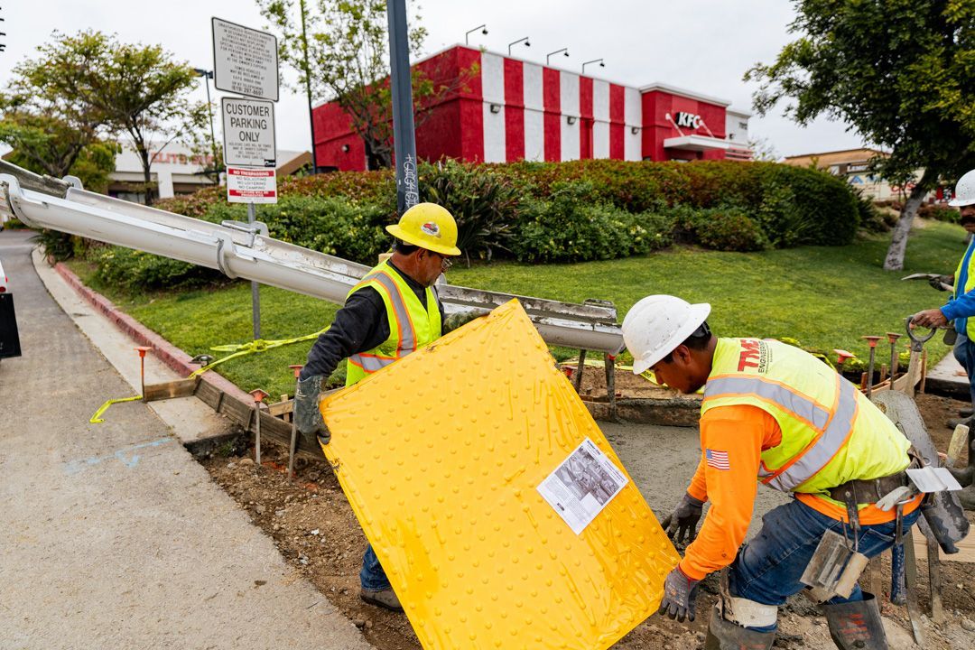 Two construction workers are working on a sidewalk in front of a kfc restaurant.