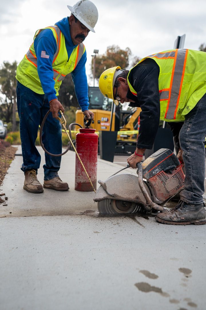 Two construction workers are working on a sidewalk and one is using a fire extinguisher.