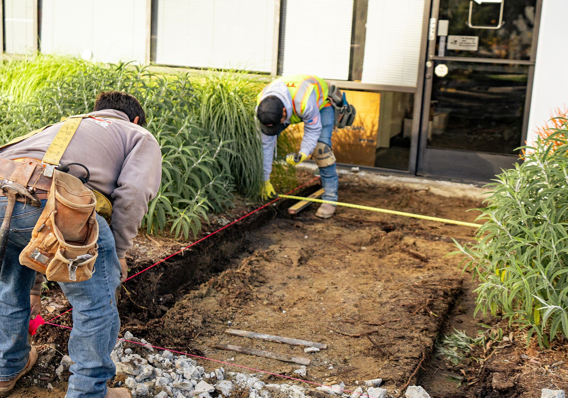Two construction workers are working on a sidewalk in front of a building.