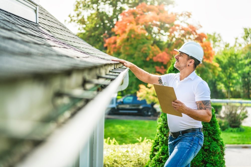 A person in a hard hat examines a gutter on a house while holding a clipboard. The sky is blue, with fall foliage.