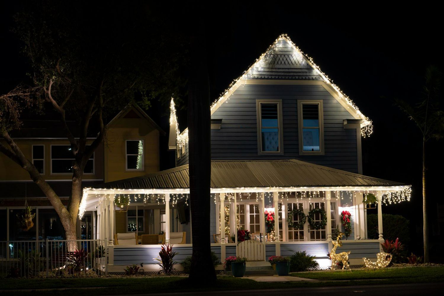A blue house at night, decorated with Christmas lights. A porch and rooftop are illuminated, with a Christmas reindeer display on the lawn.
