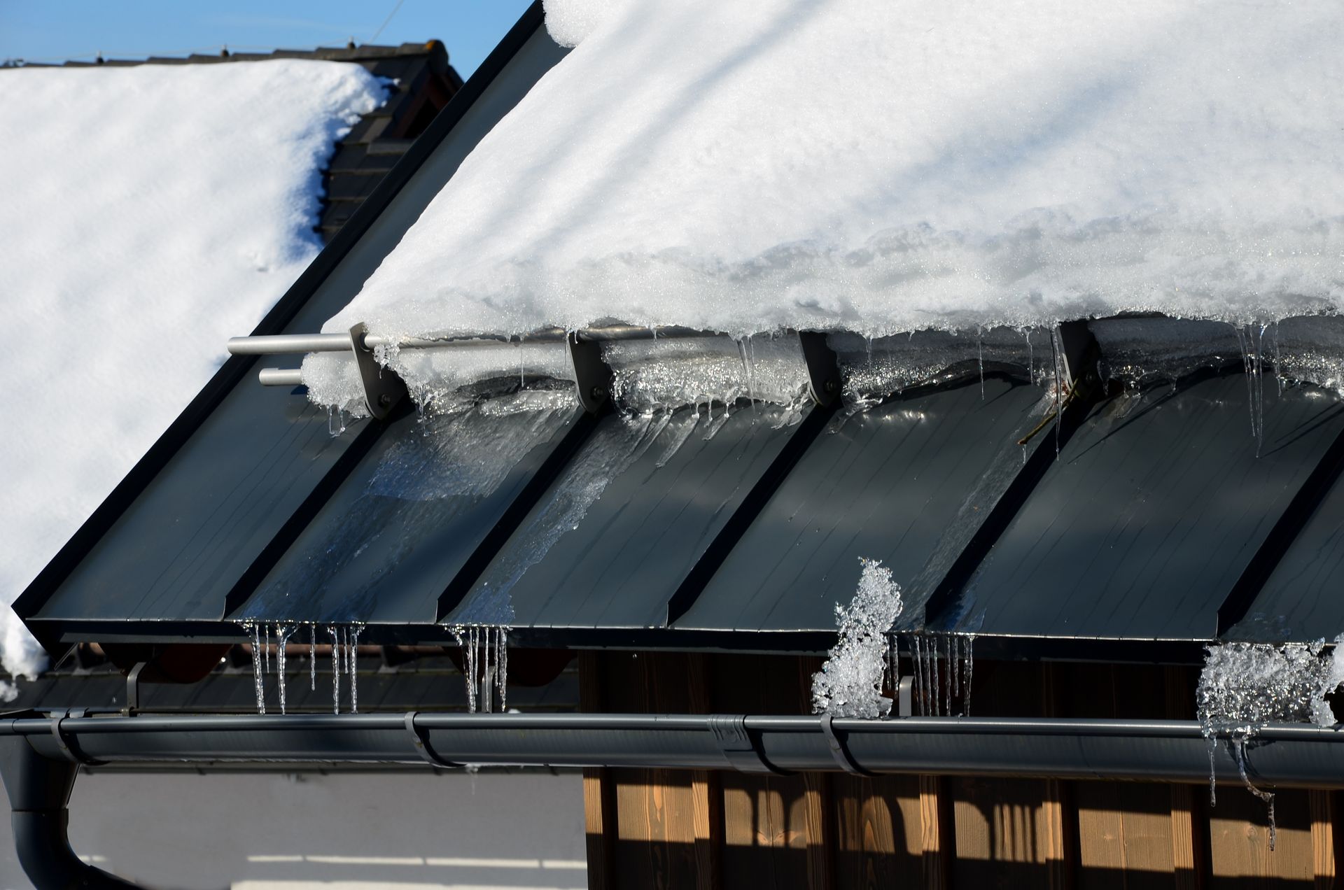 Snow and ice on a dark roof with icicles hanging from the gutter on a sunny day.