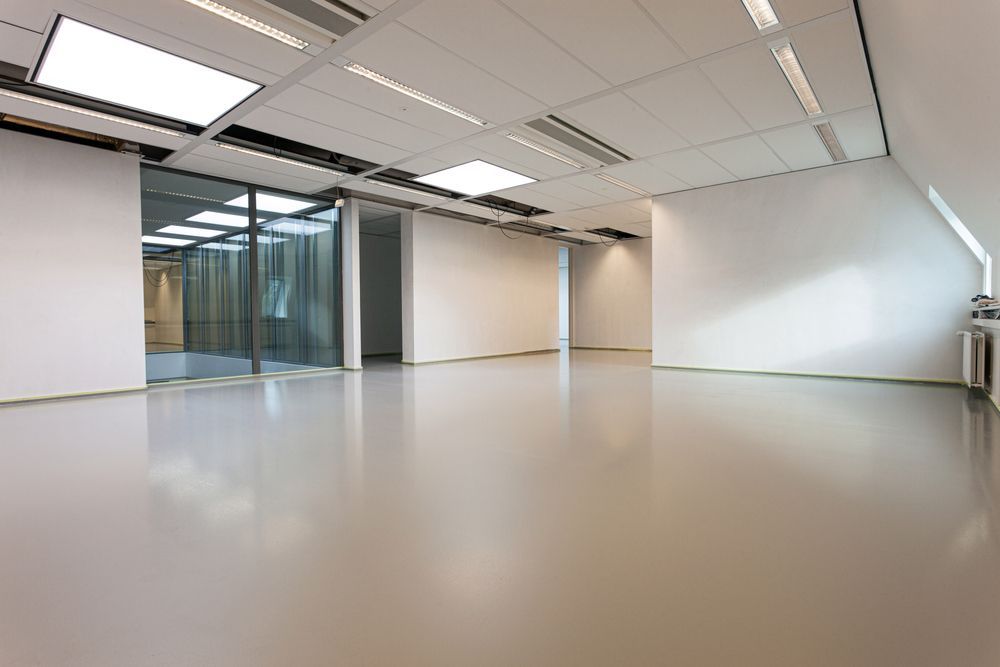 Empty office space with glossy, light-colored floor, white walls, and recessed ceiling lights. A glass wall section reveals another room.