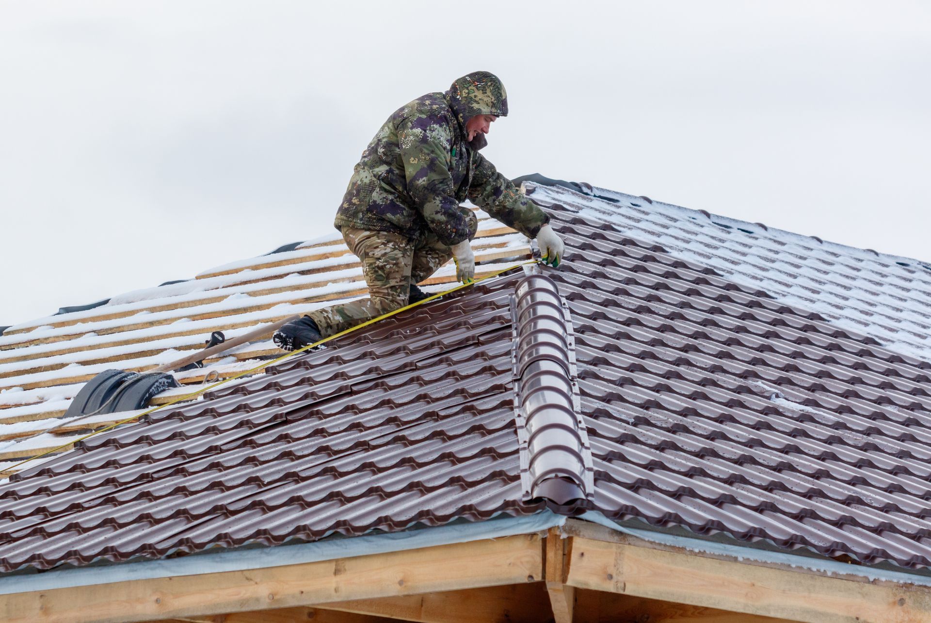 Roofer in camouflage working on a brown metal roof on a cloudy day.
