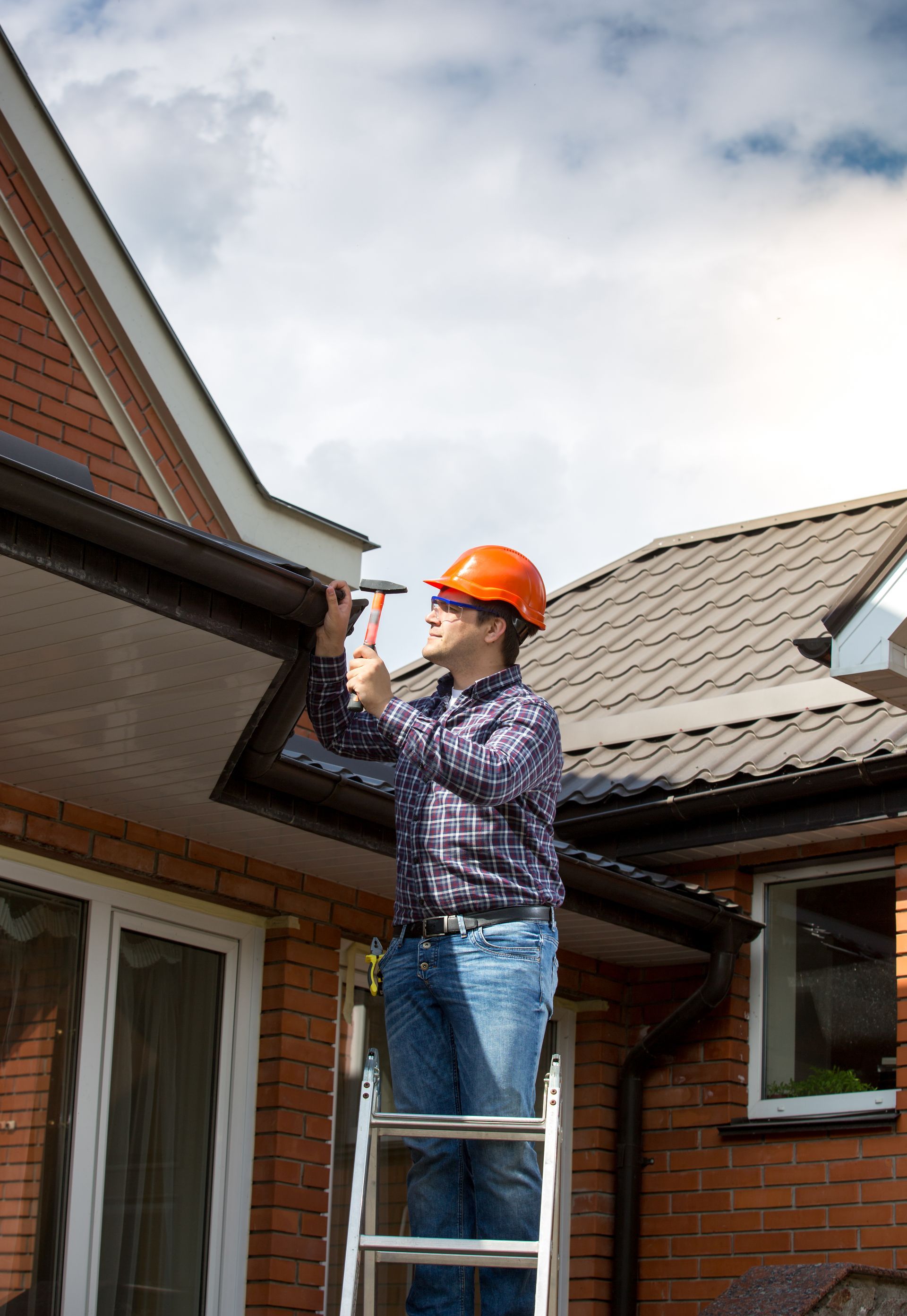 A man is standing on a ladder fixing a gutter on the side of a house.