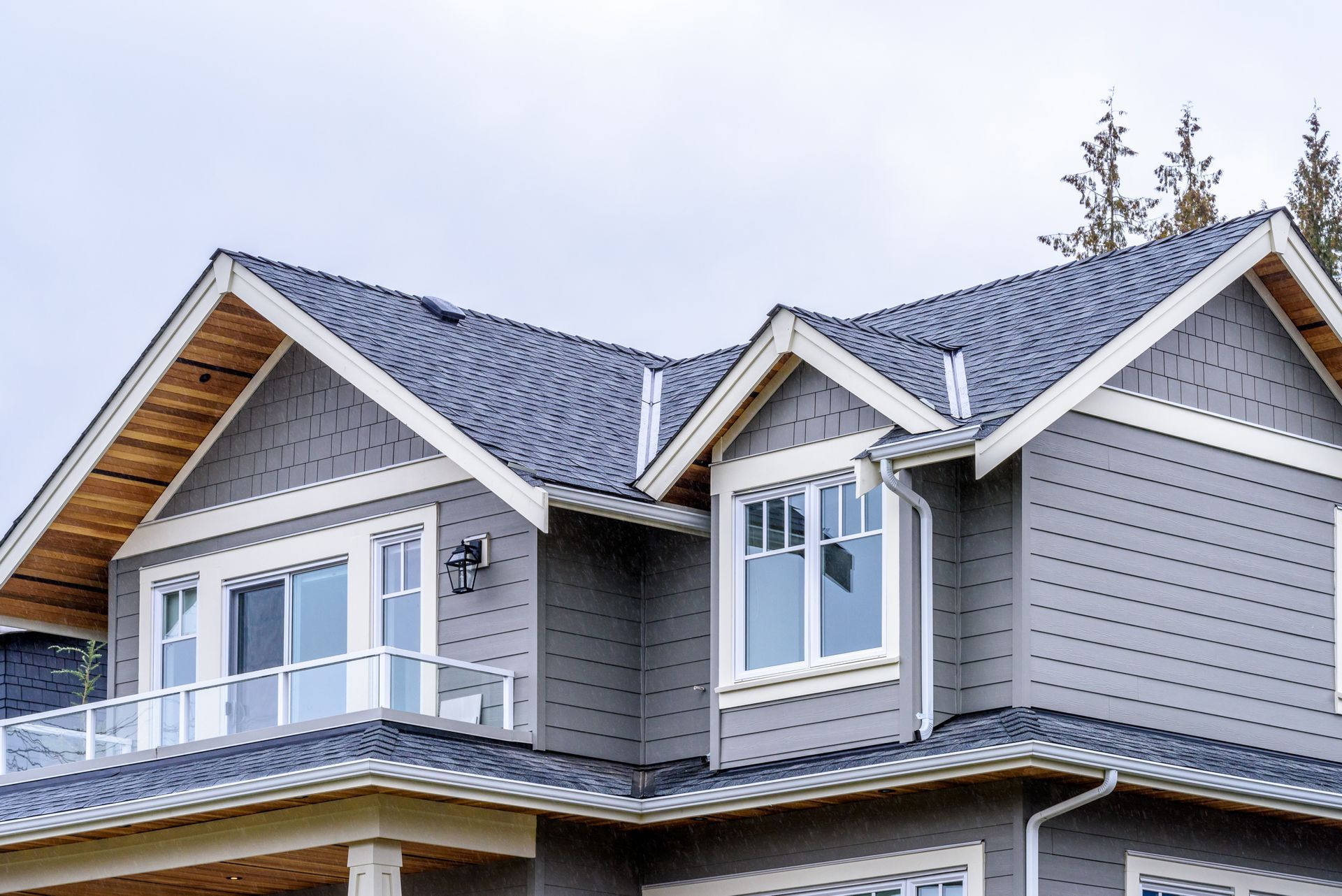 Grey house with white trim, a balcony, and a dark blue roof against a cloudy sky.