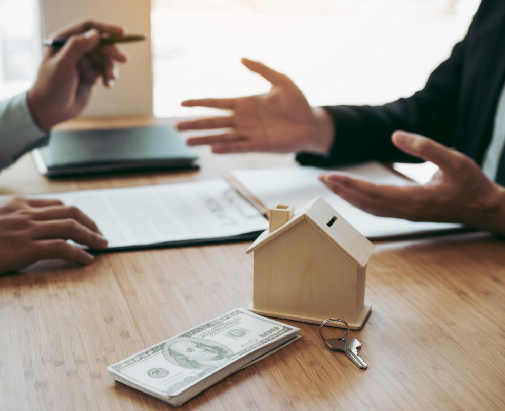 Two men are sitting at a table with a model house , money , and keys.