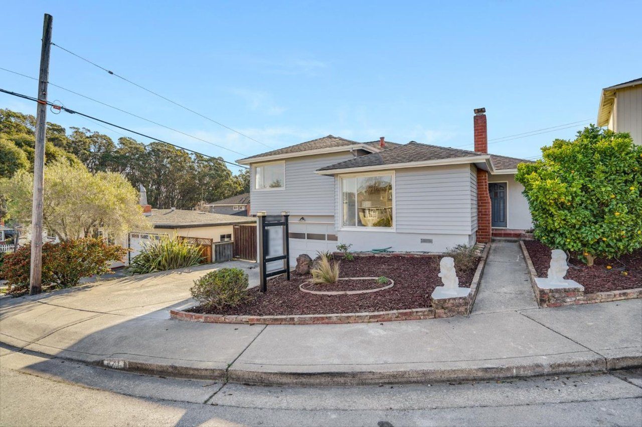 A small white house is for sale on the corner of a street.