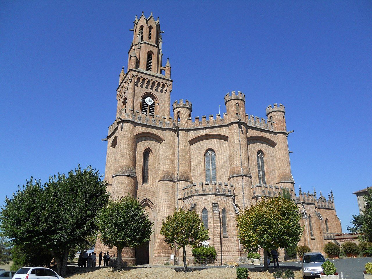 Brick church with clock tower, turrets, and arched windows under a clear blue sky, with trees in front.
