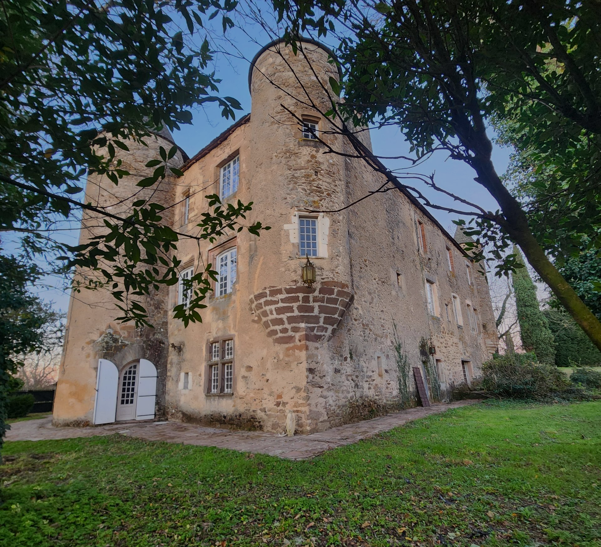 Un château historique en pierre, avec deux tours arrondies et des portes cintrées blanches, encadré d'arbres sur une pelouse verdoyante sous un ciel bleu.