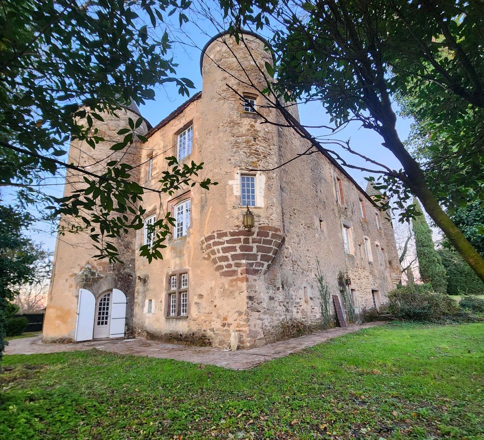 Un vieux château en pierre avec deux tours rondes, des portes blanches et des fenêtres cintrées, entouré d'arbres et d'une cour gazonnée.
