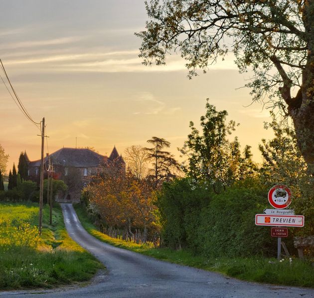 Au coucher du soleil, une route étroite mène à un château français, avec un panneau indiquant le village au premier plan.