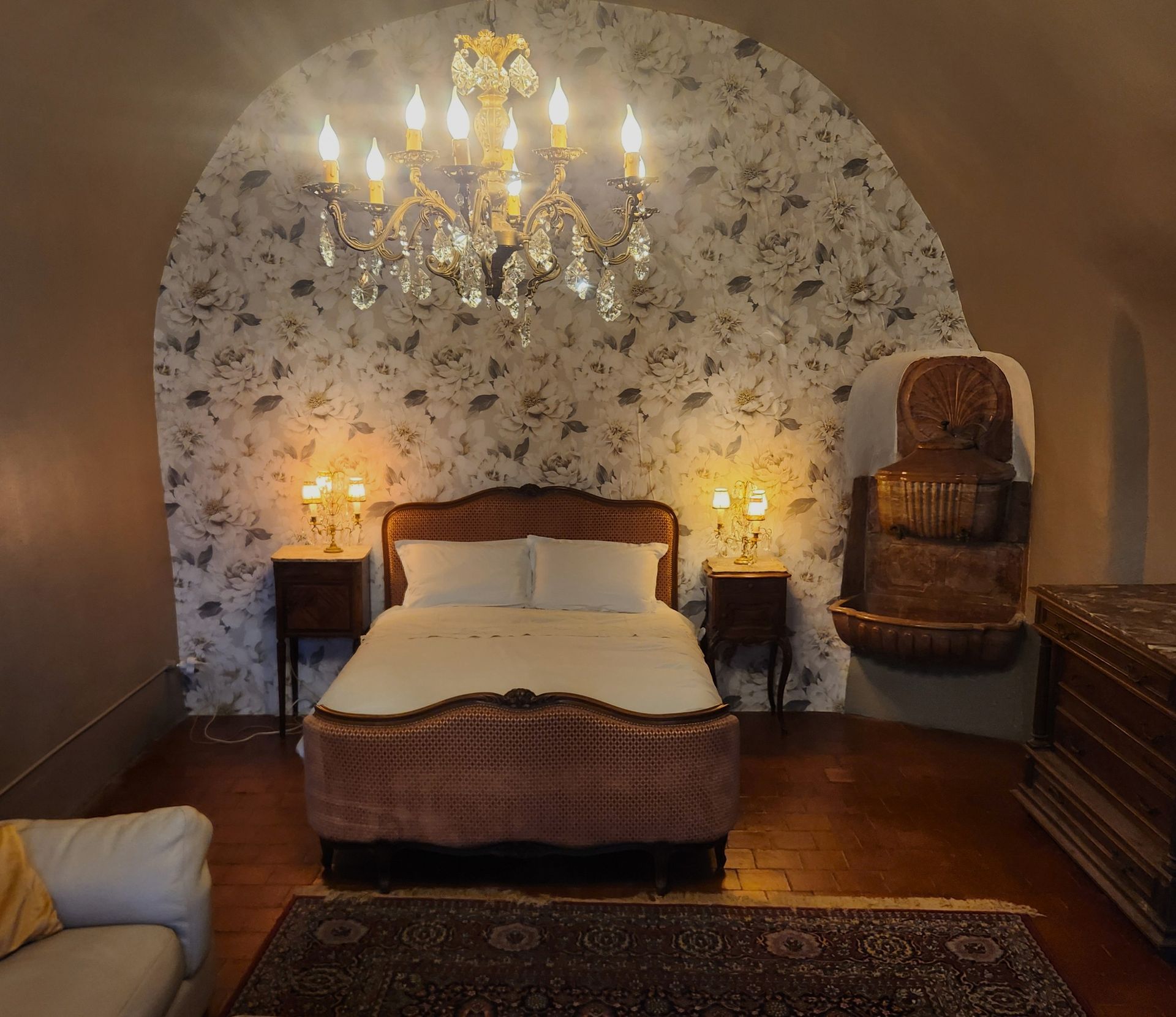 Chambre avec mur voûté, papier peint à motifs, lustre en cristal, lit avec tables de chevet et meubles en osier.