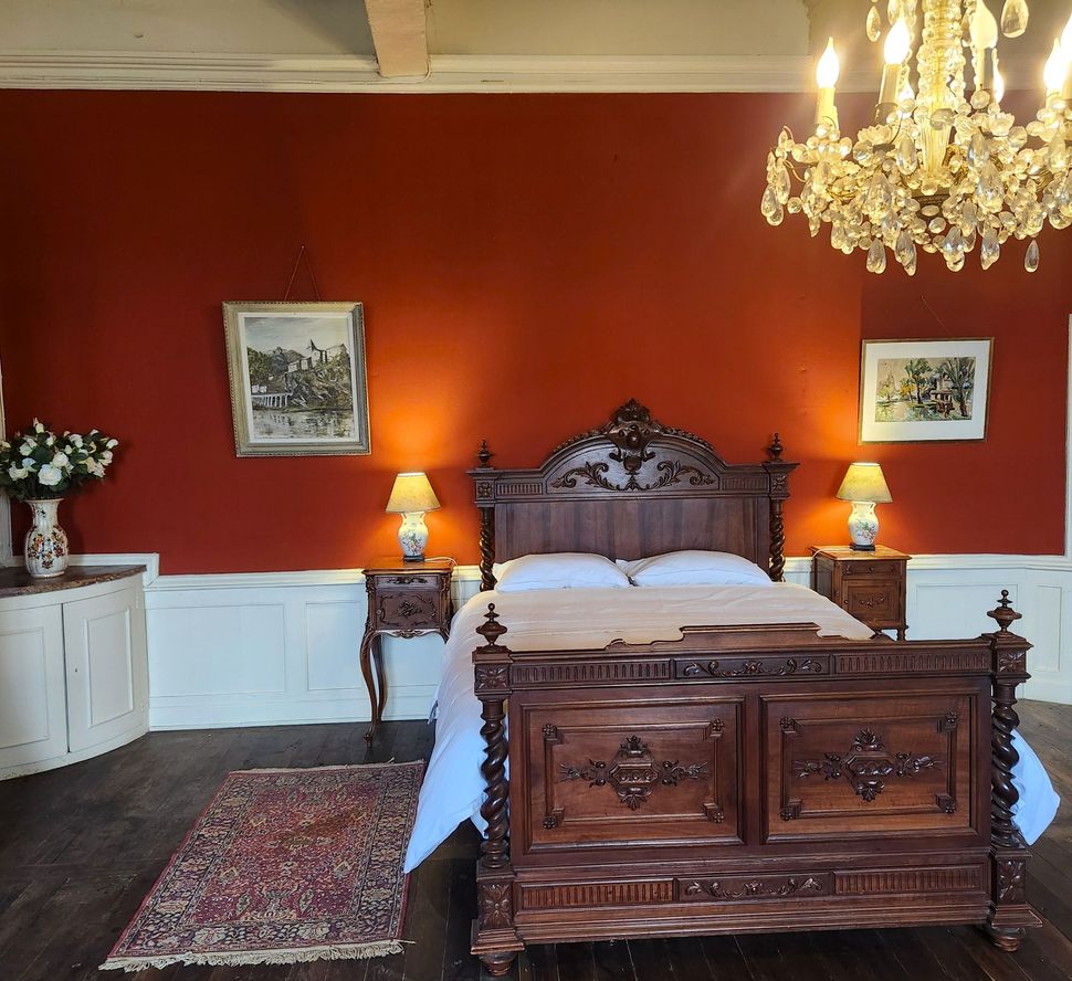 Dans une chambre aux murs couleur terre cuite, aux lambris blancs, au lustre en cristal et aux deux tapis, se dresse un lit en bois sculpté.