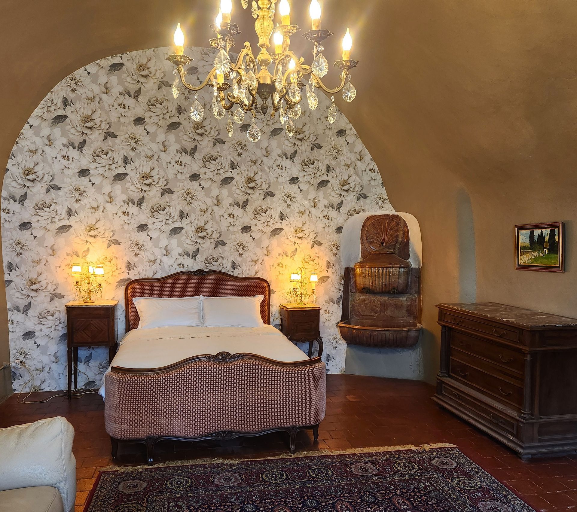 Chambre avec mur voûté, papier peint à motifs, lustre en cristal, lit avec tables de chevet et meubles en osier.