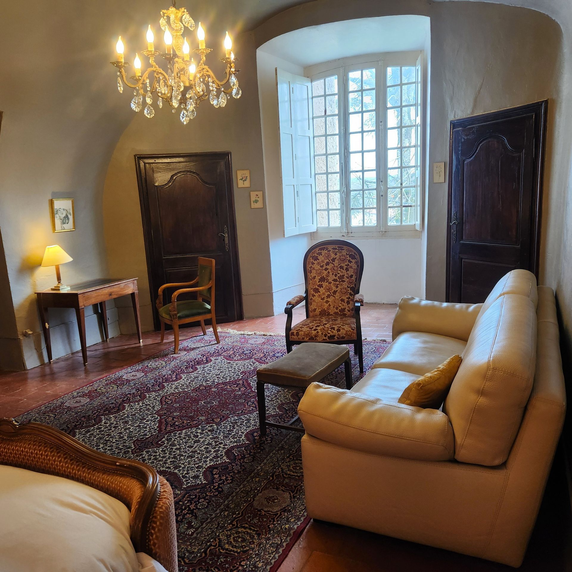 Une grande pièce voûtée avec un lustre, un tapis à motifs, un canapé beige, des chaises en bois et une grande fenêtre.