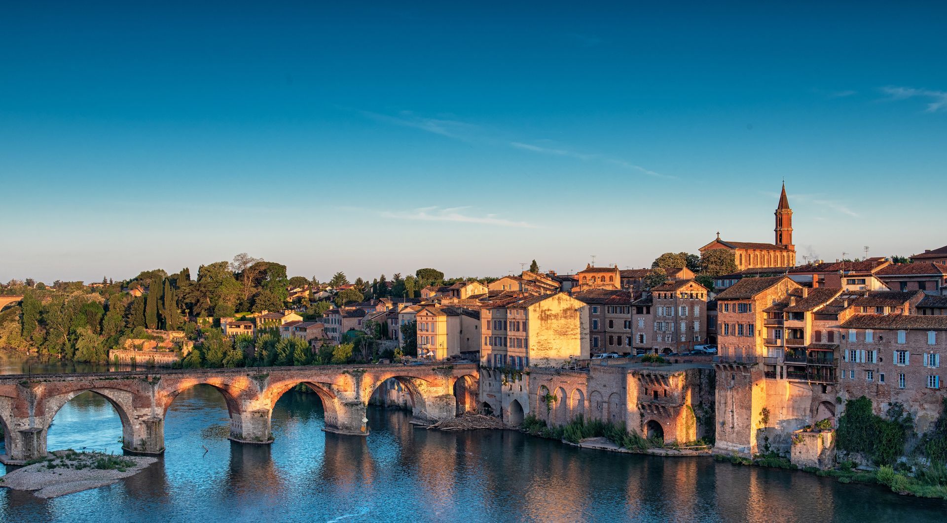 Stone bridge over a river, with a town of buildings and a church with a tall steeple in the background; sunny day.