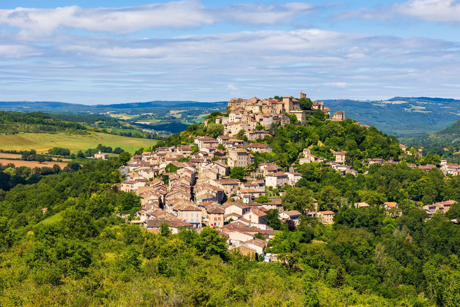 Stone village atop a hill, surrounded by green trees and fields, under a blue sky.