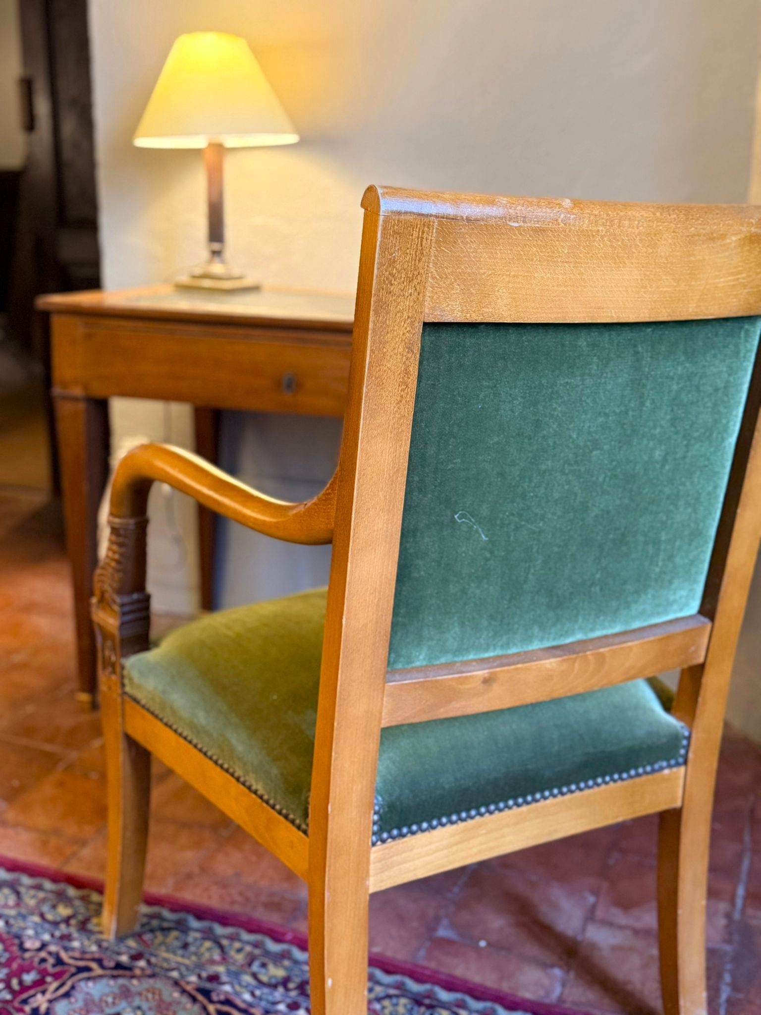 Un fauteuil en bois recouvert de velours vert se trouve devant un bureau avec une lampe, posé sur un tapis à motifs.