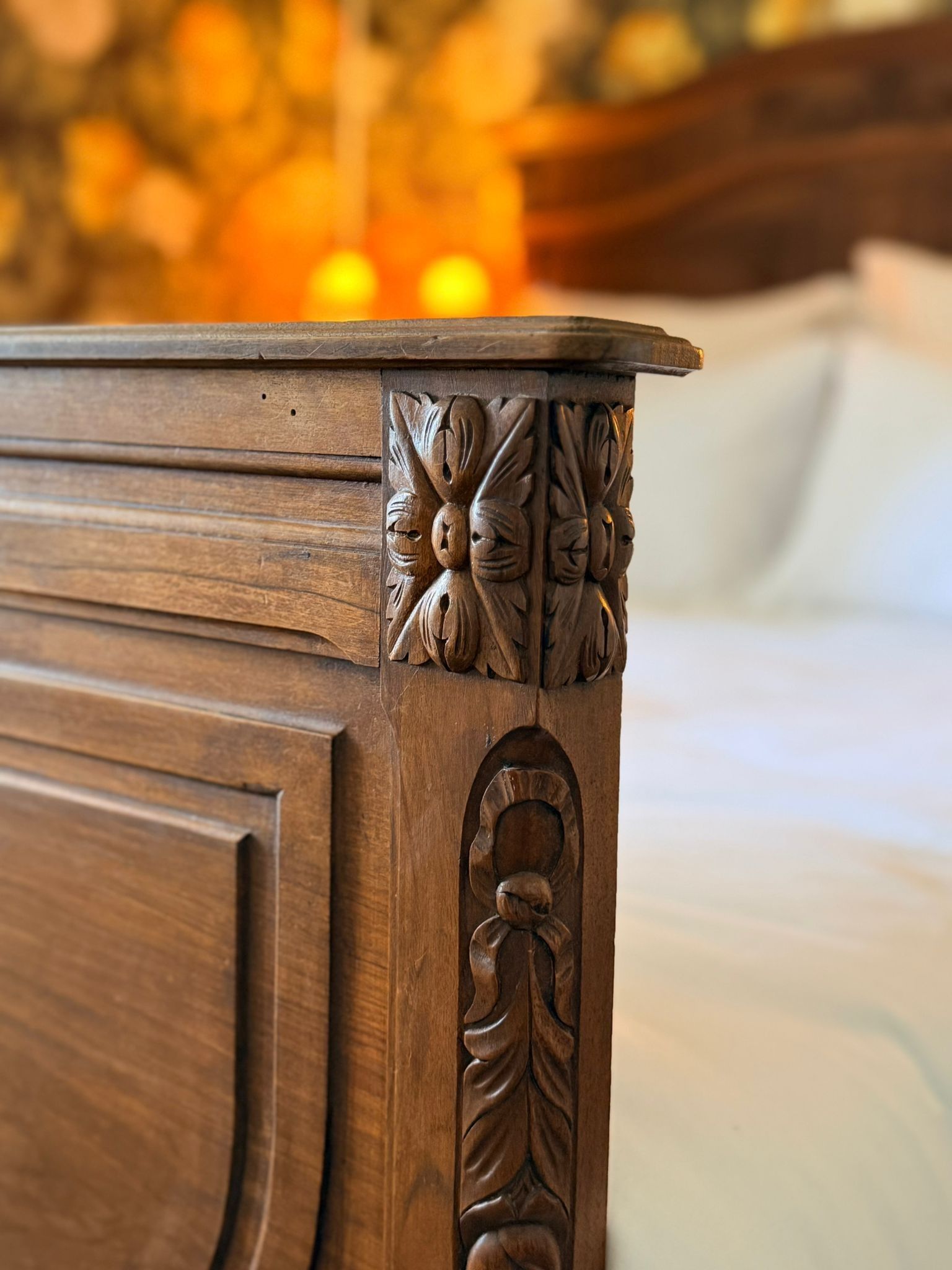 Gros plan sur un montant de lit en bois sculpté aux détails floraux et végétaux complexes, dans une chambre floue à l'éclairage chaleureux.