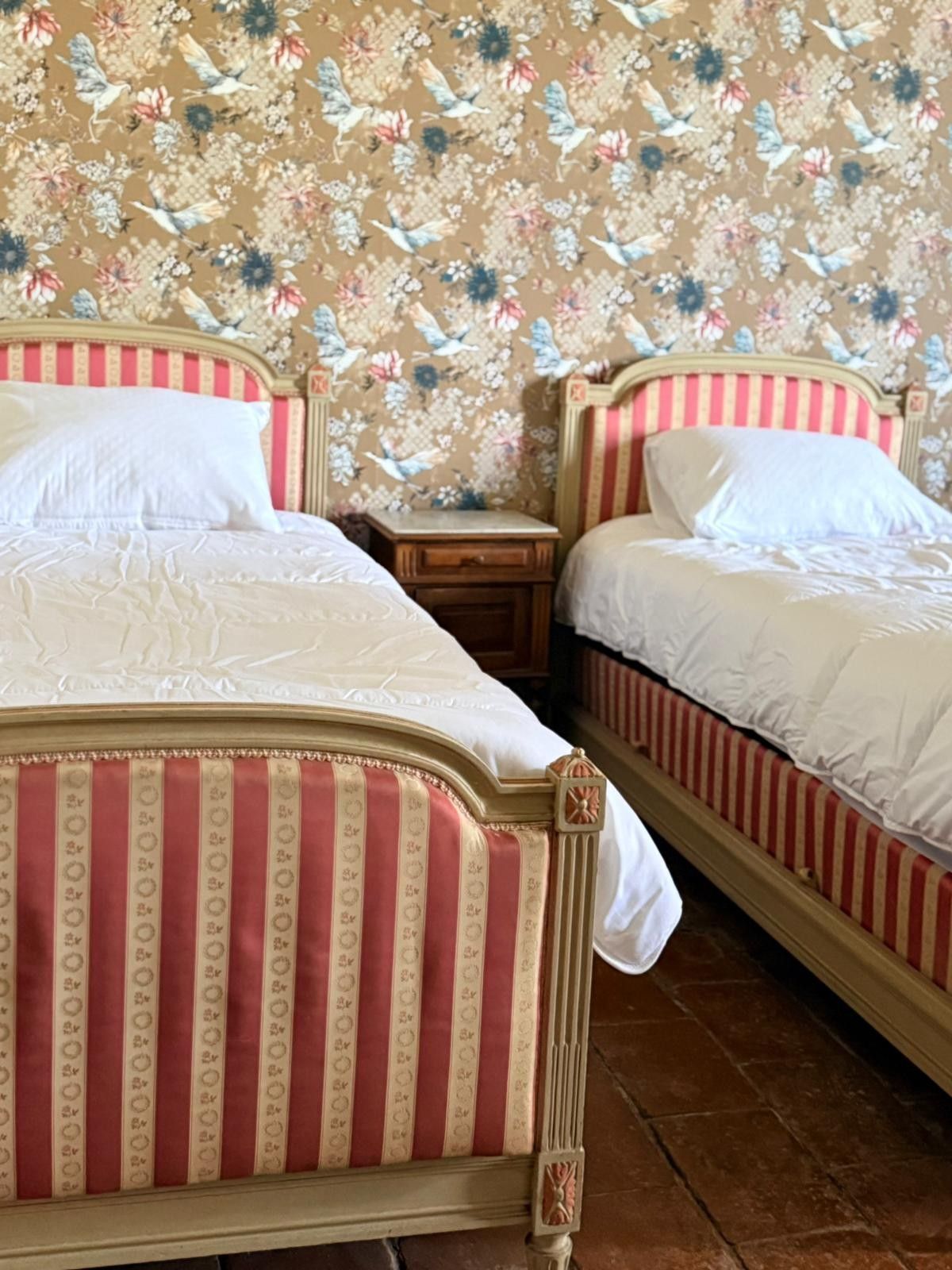 Deux lits jumeaux avec têtes de lit et cadres à rayures rouges, linge de lit blanc et une table de chevet contre un mur tapissé de papier peint à motifs floraux.