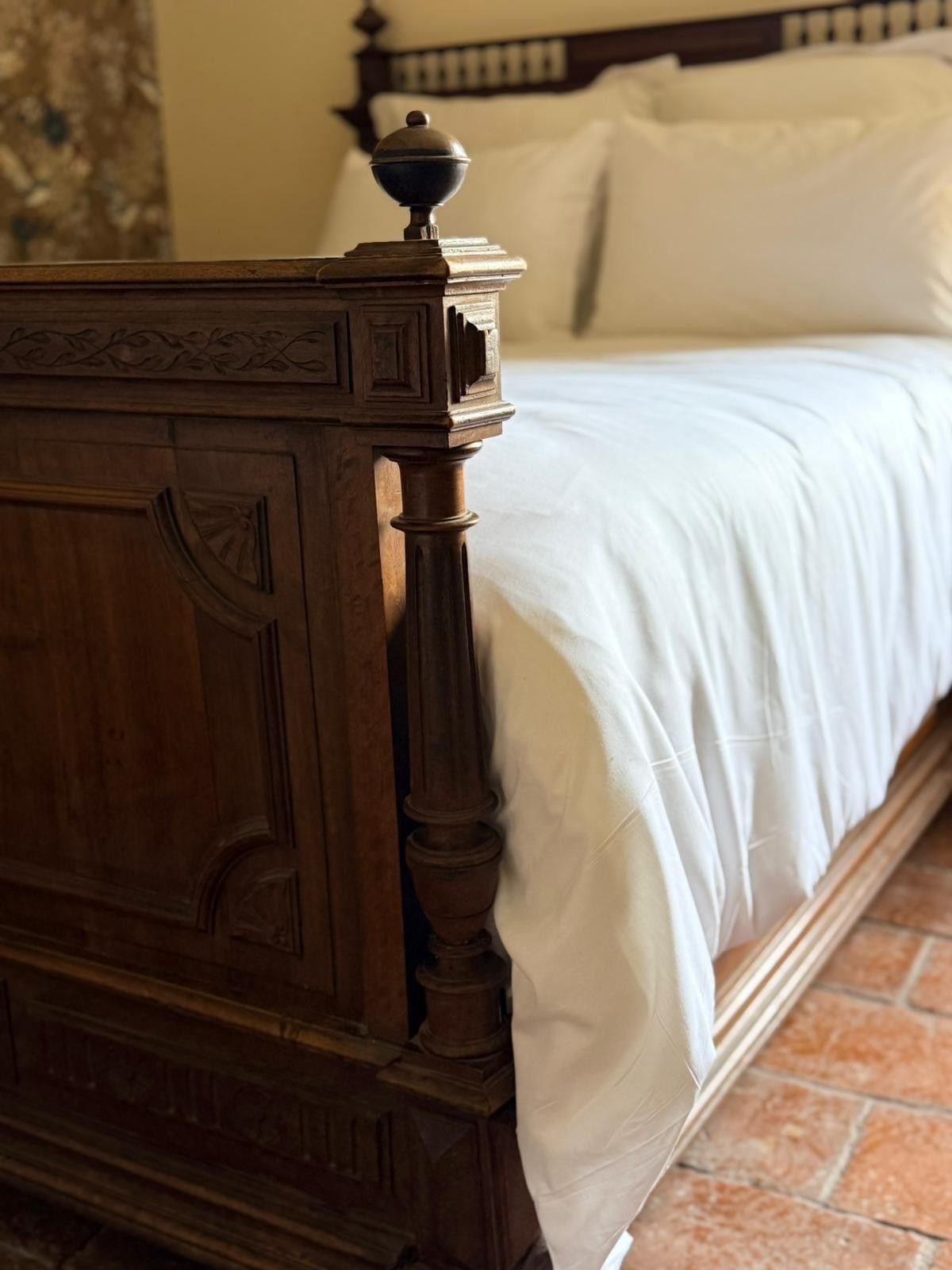 Gros plan sur un cadre de lit en bois sculpté avec une couette et des oreillers blancs sur un sol en terre cuite.